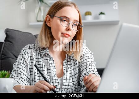 Confident blonde woman in glasses working using laptop while making notes in office interior. Student studying e learning at home. Stock Photo