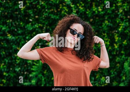Playful young woman wearing sunglasses flexing muscles in front of ivy plants Stock Photo