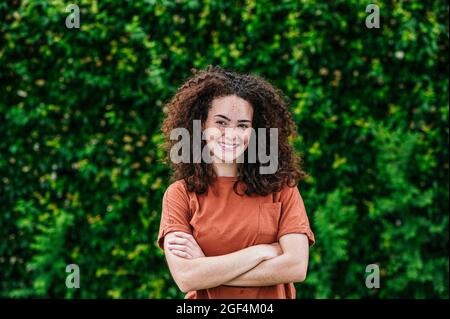 Happy young curly haired woman standing with arms crossed Stock Photo