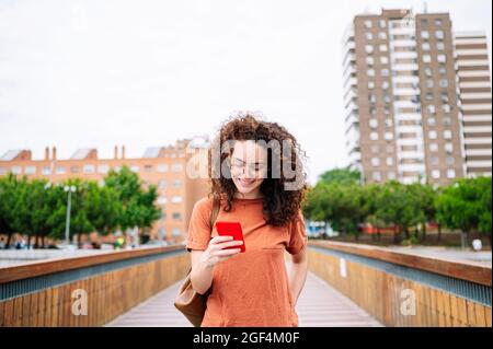 Smiling curly haired woman using smart phone while walking on bridge Stock Photo