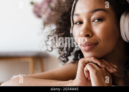 Smiling mid aged african woman listening to music with headphones while relaxing on bed at home, close up Stock Photo
