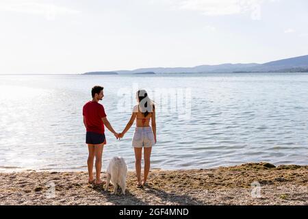 Couple holding hands while standing with dog at lakeshore Stock Photo