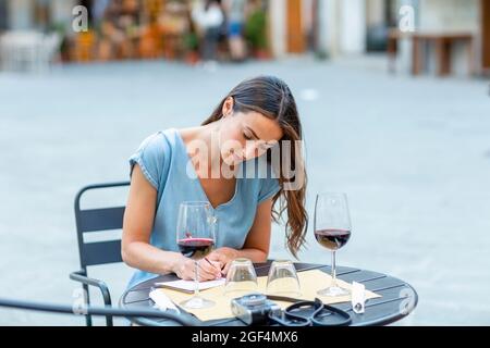 Young woman writing on postcard while sitting at sidewalk cafe Stock Photo