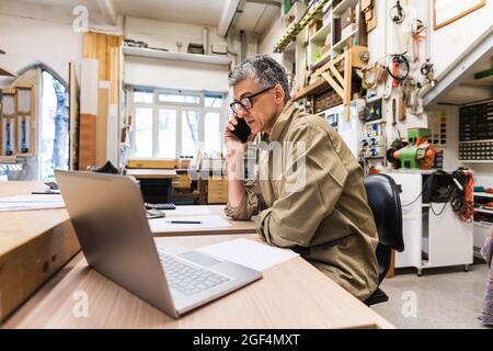 Male owner talking on mobile phone at table in workshop Stock Photo