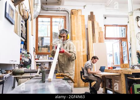 Mature male carpenter working on wood while coworker writing in workshop Stock Photo