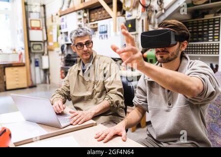 Male craftsperson using virtual reality simulator by colleague at workbench Stock Photo