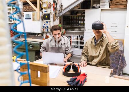 Male carpenter using virtual reality simulator while coworker using laptop in workshop Stock Photo