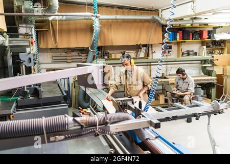 Mature carpenter working with machinery while colleague using laptop in workshop Stock Photo