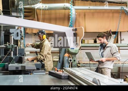 Mature craftsperson working on machinery while male colleague using laptop in workshop Stock Photo