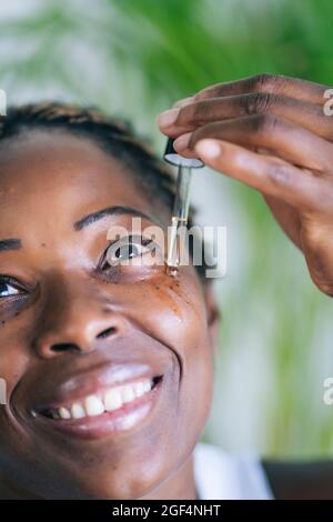 Afro woman smiling while applying face serum through dropper Stock Photo