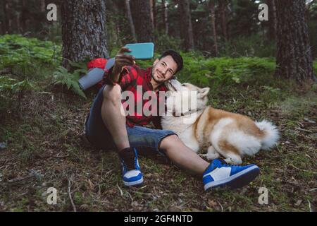 Smiling male hiker taking selfie with pet through smart phone while sitting in forest Stock Photo
