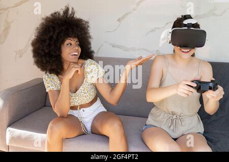 Young woman playing game while female friend gesturing sitting on sofa at home Stock Photo