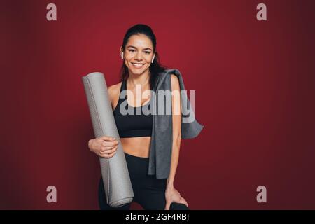 Smiling mid adult sportsperson with exercise mat and towel against maroon background Stock Photo