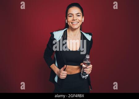 Smiling sportswoman in sports clothing holding water bottle against maroon background Stock Photo