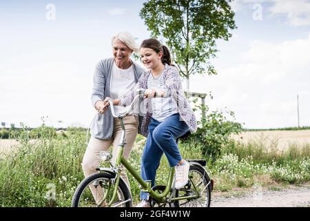 Smiling woman teaching girl riding bicycle on dirt road Stock Photo