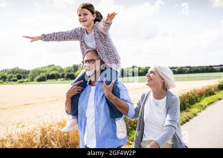Smiling girl with arms outstretched sitting on man shoulder during sunny day Stock Photo