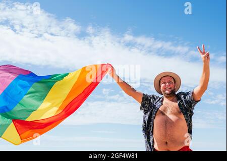 Cheerful gay man waving rainbow flag under sky on sunny day Stock Photo