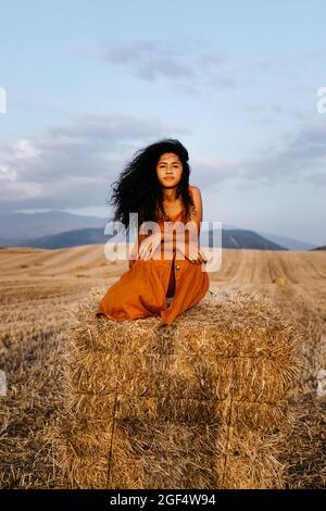Young woman crouching on hay bale during sunset Stock Photo