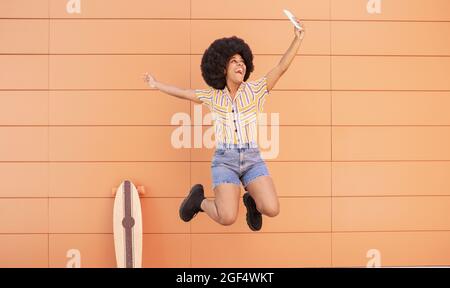 Happy woman taking selfie through smart phone while jumping in front of wall Stock Photo