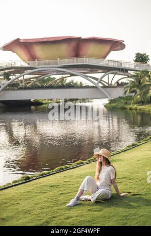 Young beautiful blonde woman wearing white casual clothes, straw hat and sneakers sitting on the grass in the park. Trendy casual spring outfit. Stock Photo