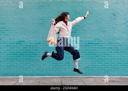 Excited young woman jumping while holding mesh bag by turquoise brick wall Stock Photo