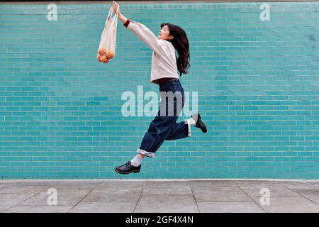Carefree young woman with fruits in mesh bag jumping by brick wall Stock Photo