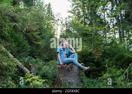 Woman resting in forest in park. dressed in denim suit and boots. Young caucasian woman sitting on tree stump surrounded by forest. Stock Photo