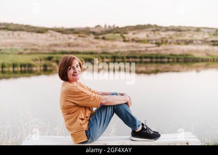 Senior woman sitting on bench by lake Stock Photo