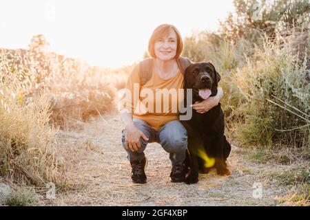 Senior woman embracing Black Labrador while crouching on trail Stock Photo