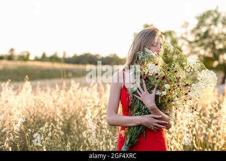 Young blond woman smelling wild flowers bouquet in field Stock Photo