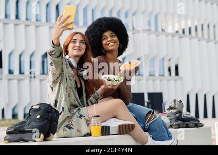 Young redhead woman taking selfie while eating with female friend Stock Photo