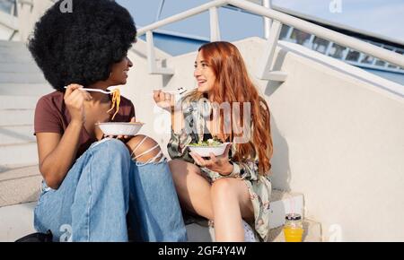 Female friends eating while sitting on steps Stock Photo