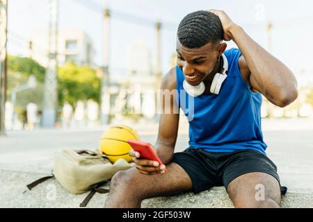 Young man with hand behind head smiling while using smart phone at basketball court Stock Photo