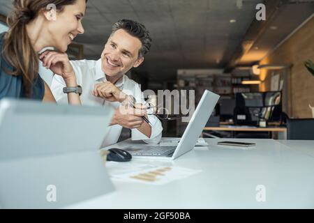 Male and female colleagues smiling while discussing over laptop at workplace Stock Photo