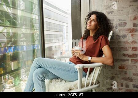 Thoughtful female professional with coffee cup sitting on chair by window Stock Photo