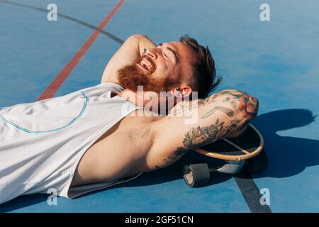 Happy young man with hands behind head resting with skateboard at basketball court on sunny day Stock Photo