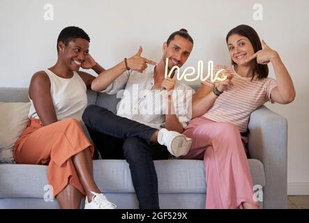 Happy male and female coworkers pointing at Hello sign illuminated text while sitting on sofa Stock Photo