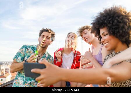 Young male and female friends blowing bubbles while taking selfie on rooftop
