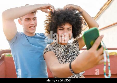Cheerful young couple taking selfie through smart phone while playing with hair on rooftop Stock Photo