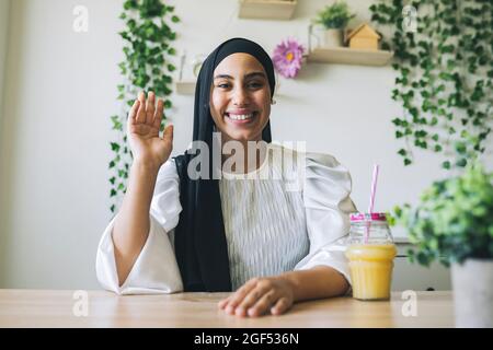 Smiling female professional waving while sitting at table in living room Stock Photo