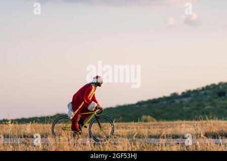 Young man in Santa Claus costume riding bicycle on road during sunset Stock Photo