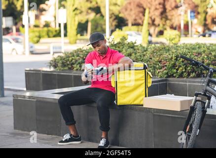 Delivery man text messaging through smart phone while sitting on bench by backpack Stock Photo