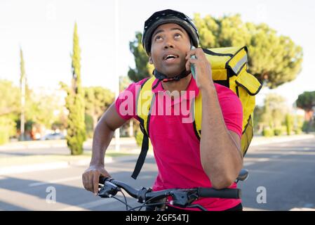 Male delivery person looking up talking on mobile phone looking up while leaning on bicycle Stock Photo