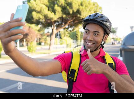 Smiling male delivery person gesturing thumbs up while taking selfie through smart phone Stock Photo