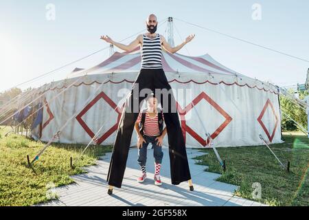 Male clown standing with performer on stilts in front of circus tent Stock Photo