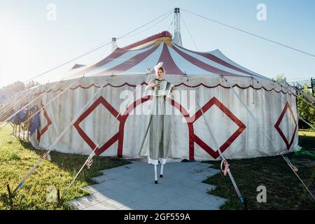 Female circus performer playing accordion while standing on stilts in front of circus tent Stock Photo