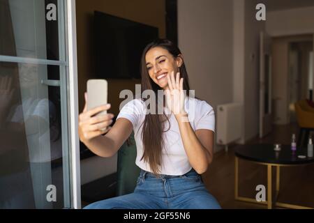 Smiling young woman waving hand during video call through smart phone Stock Photo
