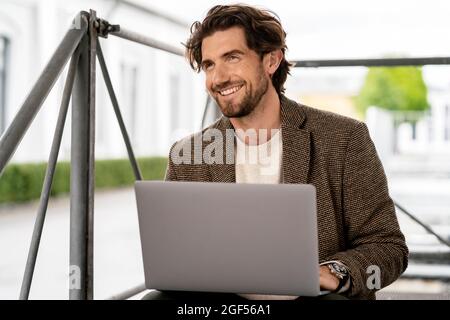 Smiling businessman looking away while sitting with laptop Stock Photo