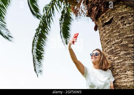 Young woman with sunglasses taking selfie through smart phone near palm tree Stock Photo