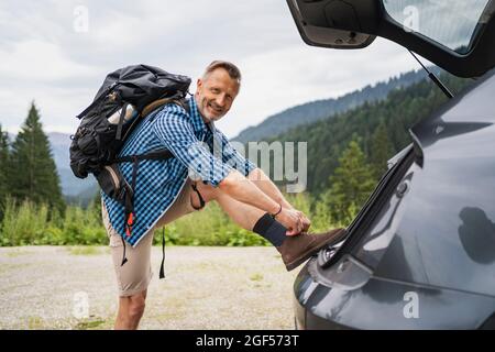 Male backpacker tying shoelace while leaning on car trunk Stock Photo